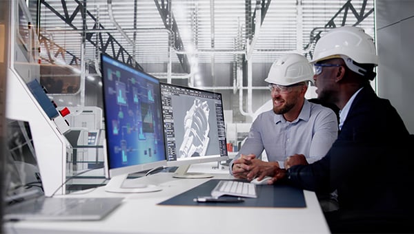 Two engineers wearing hard hats review technical designs on computer monitors in an industrial setting