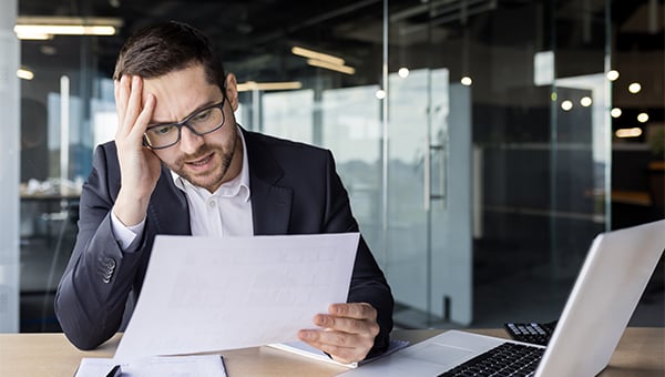 A man in an office looks stressed while reviewing a document, holding his head with one hand beside a laptop