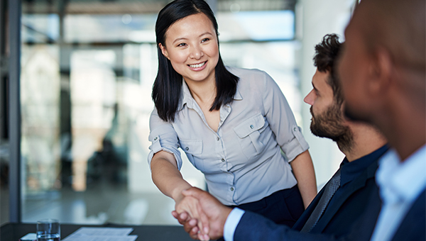 Woman smiling and shaking hands with a seated colleague during a meeting in a modern office.
