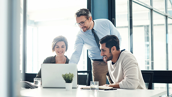 Three colleagues in a modern office gathered around a laptop, smiling and collaborating while one points at the screen.