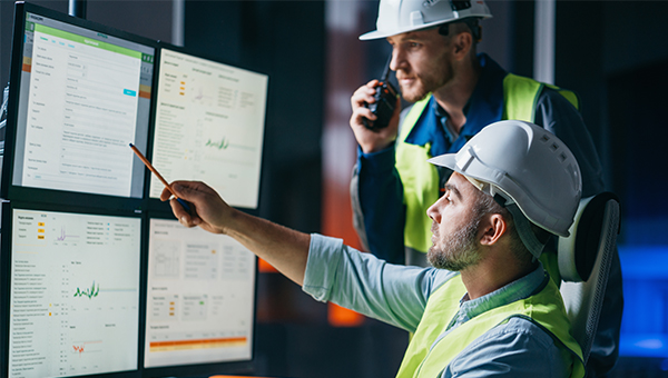 Two workers in safety vests and hard hats monitoring data on multiple screens, with one pointing at the display while the other speaks into a radio.