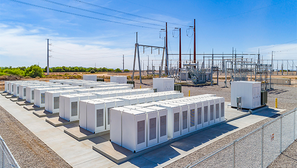 Electrical substation with multiple white battery storage units arranged in rows, surrounded by transformers, power lines, and fencing under a clear blue sky.