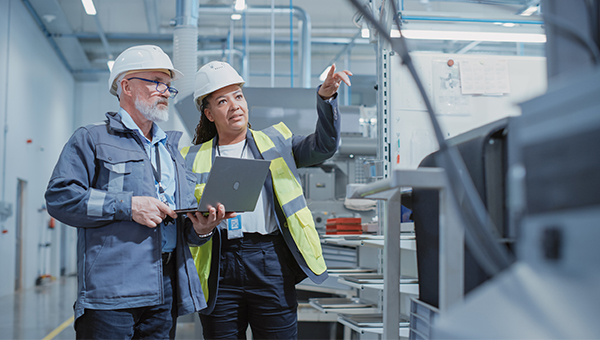 Two engineers in hard hats and safety gear review data on a laptop while one points toward industrial equipment inside a facility.