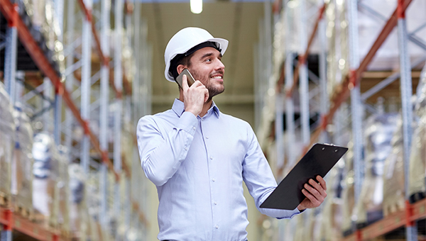 Warehouse worker wearing a hard hat talks on a phone while holding a clipboard, standing between tall storage racks.