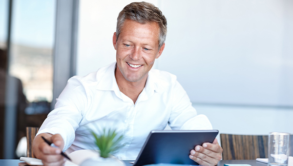 Smiling man in a white shirt sitting at a desk, holding a tablet and writing on paper with a pen in a bright office setting.