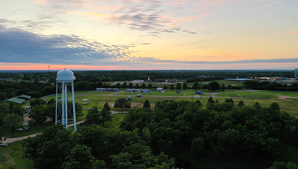 Aerial view of a white water tower surrounded by trees, open fields, and small buildings at sunset.