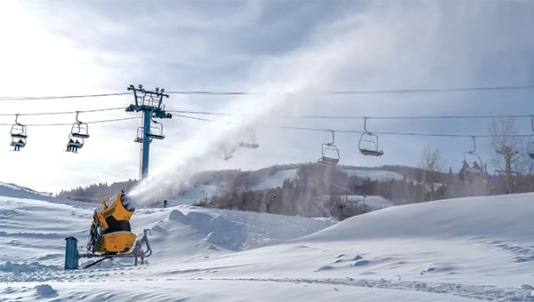 Snowmaking machine spraying snow on a ski slope with empty chairlift chairs overhead.