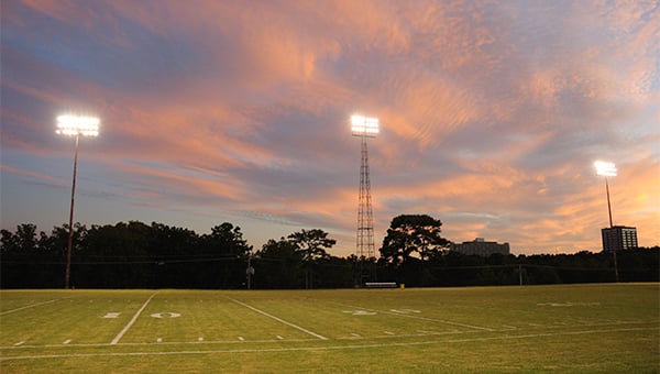 Football field with yard lines and tall stadium lights lit against a colorful sunset sky.