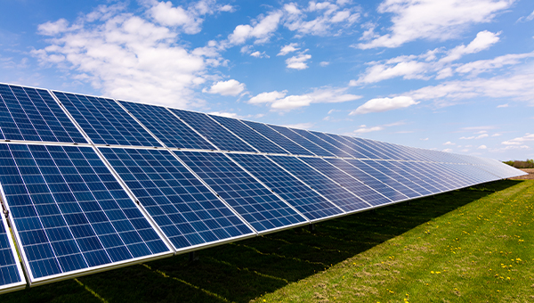 Rows of solar panels installed across a grassy field under a blue sky with scattered clouds.