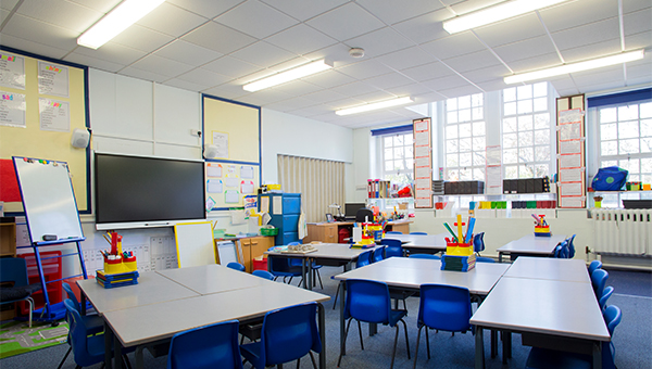 Empty classroom with tables, blue chairs, teaching supplies, and large windows letting in daylight.