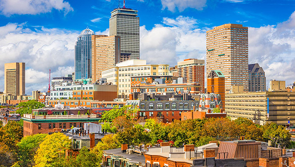 City skyline with a mix of modern high-rise buildings and older brick structures under a partly cloudy blue sky, with trees and rooftops in the foreground.