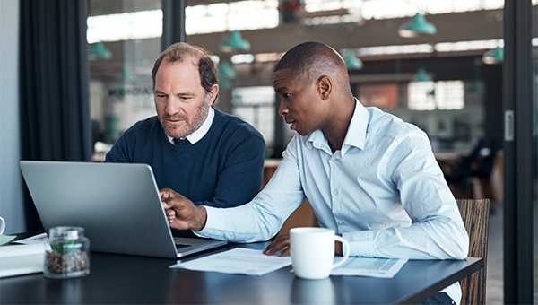 Two coworkers reviewing information on a laptop at a table, with documents and a coffee mug nearby in an office setting.