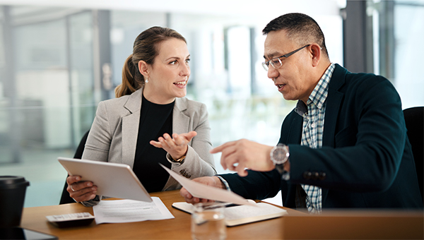 Two colleagues discuss documents at a desk, with one holding a tablet while the other points at paperwork