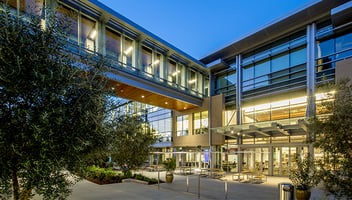 A modern building exterior at dusk with large glass windows, outdoor seating, and landscaped greenery.