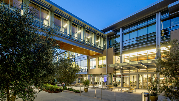 A modern building exterior at dusk with large glass windows, outdoor seating, and landscaped greenery.