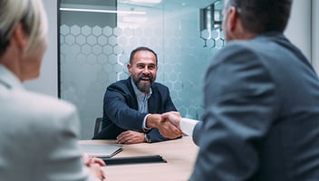 A man shakes hands with a colleague across a table during a meeting, while others sit nearby.