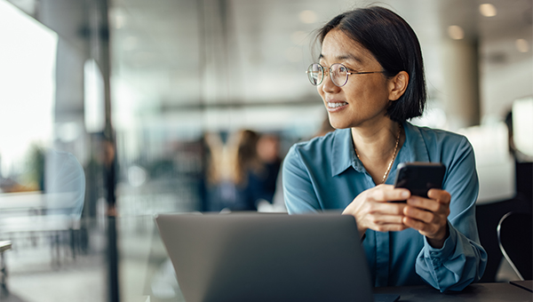 Person seated at a table in a modern office space, holding a smartphone beside an open laptop, with glass walls and other people blurred in the background.