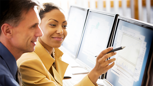 Two people seated at a desk reviewing data on dual computer monitors, with one person pointing at charts on the screen using a pen.