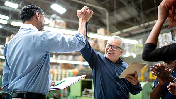 Two coworkers in a factory or warehouse celebrate with a high-five while holding a clipboard and tablet, with another colleague clapping nearby.