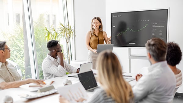 Woman presenting a stock chart on a screen to a group in a bright conference room, with attendees seated around a table using laptops and documents.