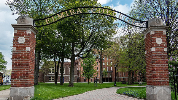 The entrance to Elmira College features a brick archway with a metal sign, leading into a tree-lined campus with green lawns and red-brick buildings.