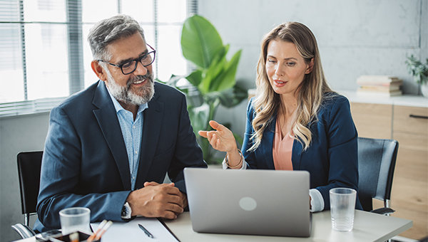 smartly dressed man and woman having a discussion at a desk in front of an open laptop in a well lit office space