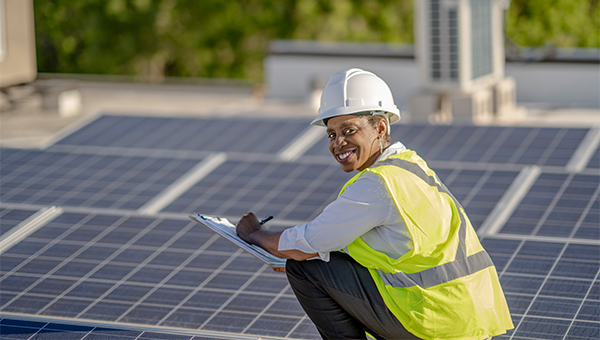 a solar panel technician wearing a heard hat and safety vest stooping by solar panels in a field taking notes on their clipboard