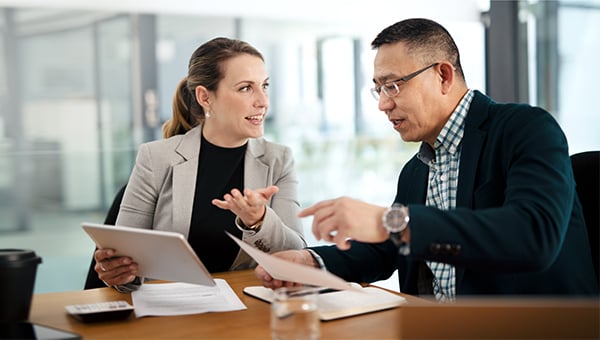 Two colleagues discuss documents at a desk, with one holding a tablet while the other points at paperwork