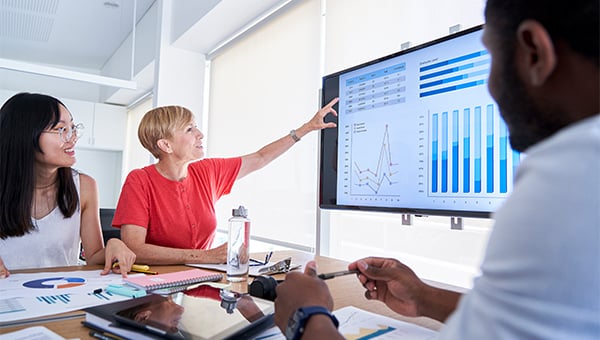Three colleagues review charts on a screen during a meeting, with one person pointing to the data while others look on