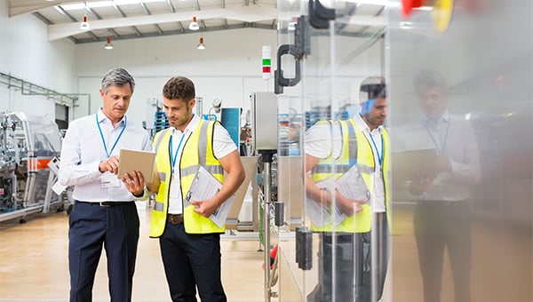 Two workers in a factory review a tablet together while standing near industrial equipment, one wearing a high-visibility vest and holding documents.