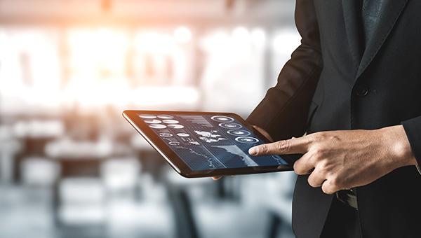 A person in a dark business suit holding a tablet and interacting with a touchscreen interface displaying charts and data, with a blurred office background.