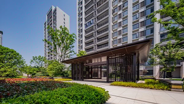 Modern apartment complex with landscaped greenery and a covered entrance pavilion between tall residential buildings.