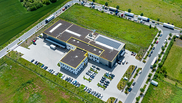 Aerial view of a commercial building with rooftop solar panels, surrounded by parking lots, roads, and green fields.