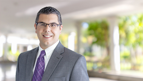 Portrait of a smiling man in glasses wearing a gray suit and purple tie, standing in a bright, blurred office or lobby setting.