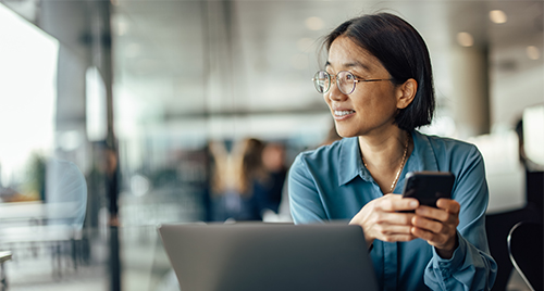 Woman wearing glasses holding a smartphone and looking to the side, seated at a table with a laptop in a modern office or café setting.