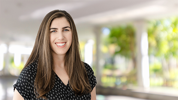 Portrait of a smiling woman with long brown hair wearing a black patterned blouse, standing in a bright, blurred indoor setting.