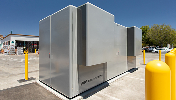 Large outdoor industrial battery storage unit (Mainspring) installed on a concrete pad, surrounded by safety bollards.