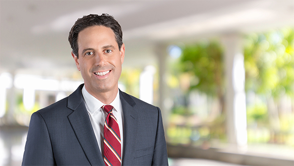 Portrait of a man in a suit and red striped tie smiling at the camera in a bright indoor setting with blurred greenery in the background.