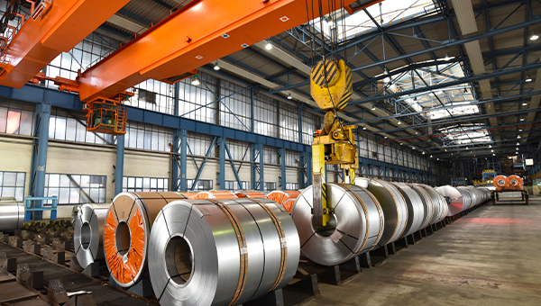 Industrial facility interior with steel coils lined up on the factory floor and an overhead crane lifting one coil.