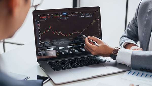 A person points to a rising financial chart on a laptop screen during a discussion with a colleague