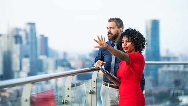 Two professionals standing on a balcony overlooking a city skyline, with one person in a red dress gesturing outward while the other holds a tablet and looks on.