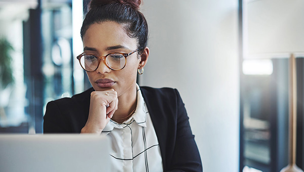 A focused professional wearing glasses rests her chin on her hand while looking at a laptop in an office setting.