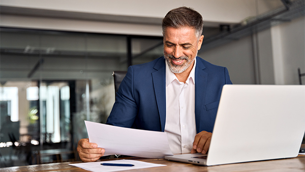 Man in a suit smiling while reviewing a document at a desk, with a laptop open beside him in an office.