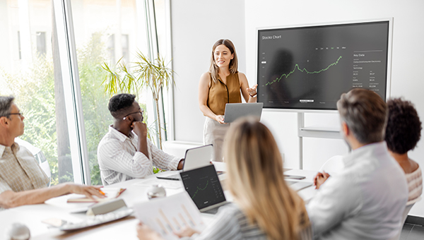 Woman presenting a stock chart on a screen to a group in a bright conference room, with attendees seated around a table using laptops and documents.