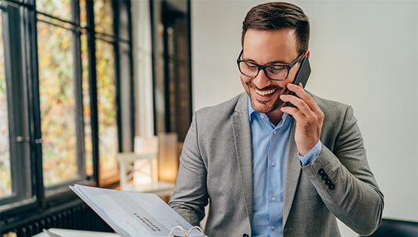 Smiling man in glasses and a gray blazer talking on a phone while holding and reading a document in a bright office setting. Smiling man in glasses and a gray blazer talking on a phone while holding and reading a document in a bright office setting.