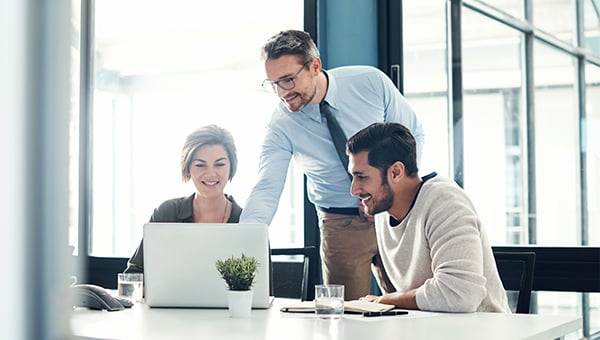 Three colleagues in a modern office gathered around a laptop, smiling and collaborating while one points at the screen. Three colleagues in a modern office gathered around a laptop, smiling and collaborating while one points at the screen.